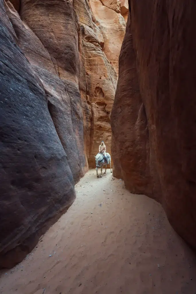 A person rides a horse through a narrow canyon with towering red rock walls.